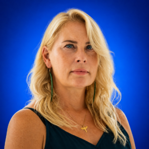 Portrait of a blonde woman with blue eyes wearing a dark teal top and green earrings against a vivid blue background.