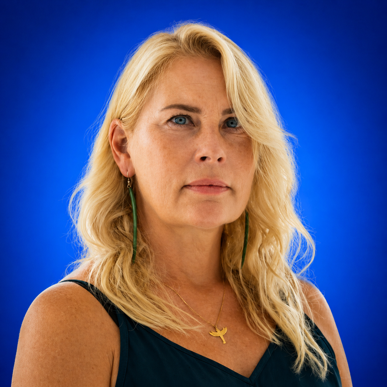 Portrait of a blonde woman with blue eyes wearing a dark teal top and green earrings against a vivid blue background.