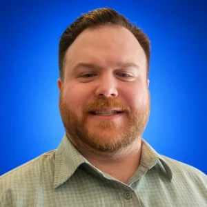Kyle Gohres Smiling man with short brown hair and a reddish beard, wearing a light green check shirt, against a blue gradient background.