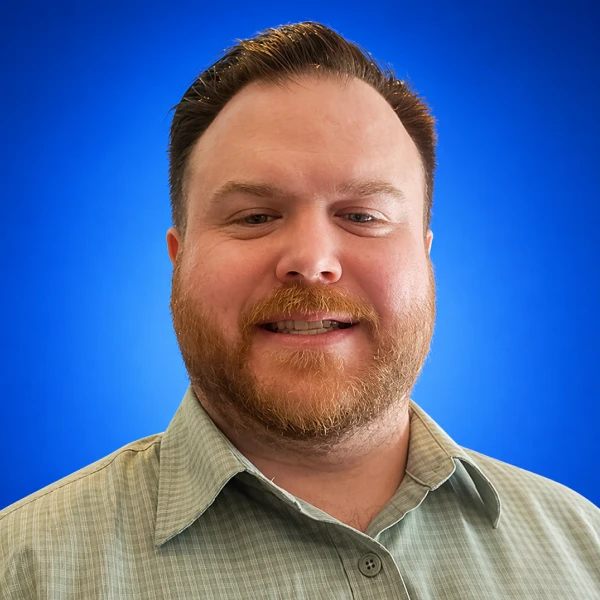 Smiling man with short brown hair and a reddish beard, wearing a light green check shirt, against a blue gradient background.