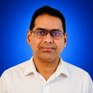 Ratan Sharmacharya Portrait of a man with glasses in a white shirt against a blue background. 1