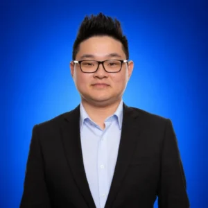 Professional headshot of a man with black hair, wearing glasses, a light blue shirt and black blazer against a blue gradient background.