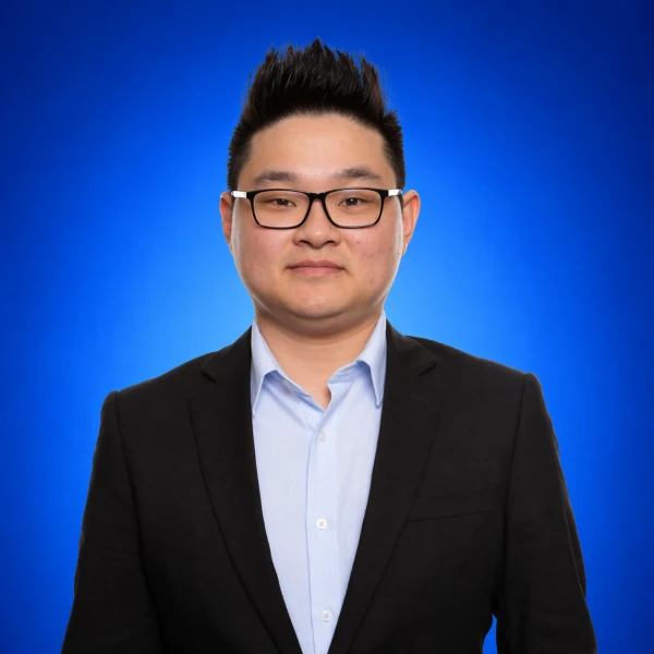 Professional headshot of a man with black hair, wearing glasses, a light blue shirt and black blazer against a blue gradient background.