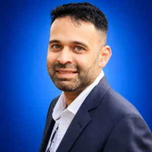 Portrait of a smiling man with short dark hair and stubble, wearing a navy blazer and white shirt against a blue backdrop.