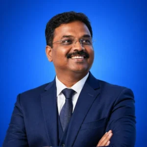 Smiling man in a navy suit, white shirt, and tie, standing against a blue backdrop with arms crossed, wearing glasses.