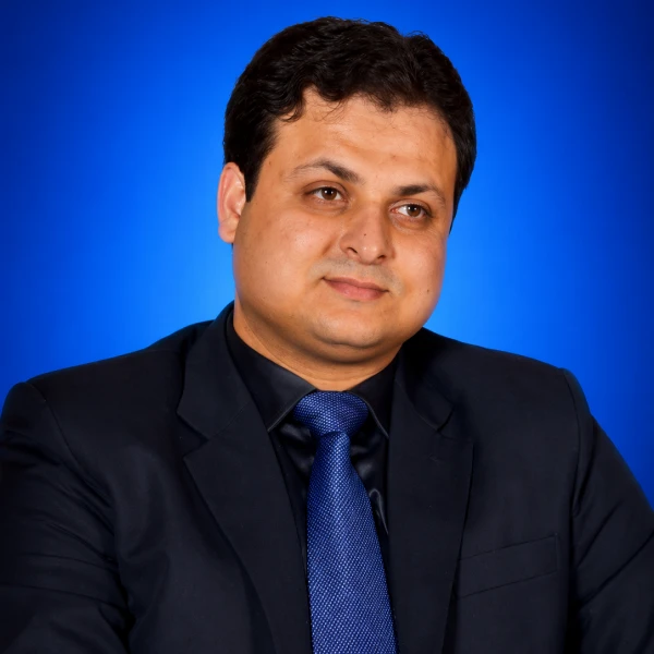 Portrait of a man in a dark suit and blue tie against a blue studio background, looking slightly to the side.