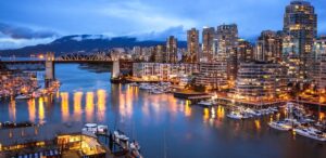 City skyline at blue hour over a marina, lights reflecting on calm water with a bridge to the left and boats docked along the shore, tall buildings rising in the background.