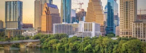 City skyline with tall modern buildings, a white multi-story structure near the river, and a tree-filled foreground with a bridge crossing the water at sunset.