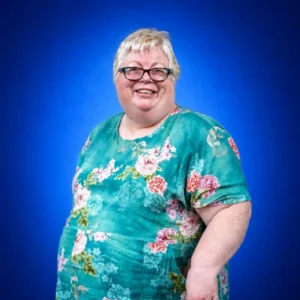 Smiling woman with short blonde hair and glasses wearing a teal floral dress against a blue background.