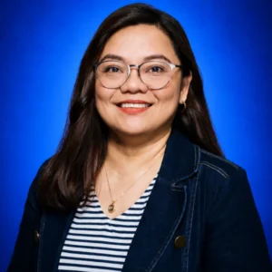 Smiling woman with glasses and long dark hair in a denim jacket against a blue studio backdrop.