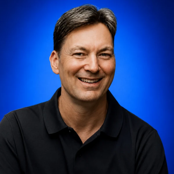 Smiling man with short gray-streaked hair wearing a black polo shirt against a blue studio backdrop