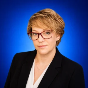 Natalia Moyseyenko Professional headshot of a woman with short blonde hair and red glasses, wearing a black blazer and white blouse against a blue background.