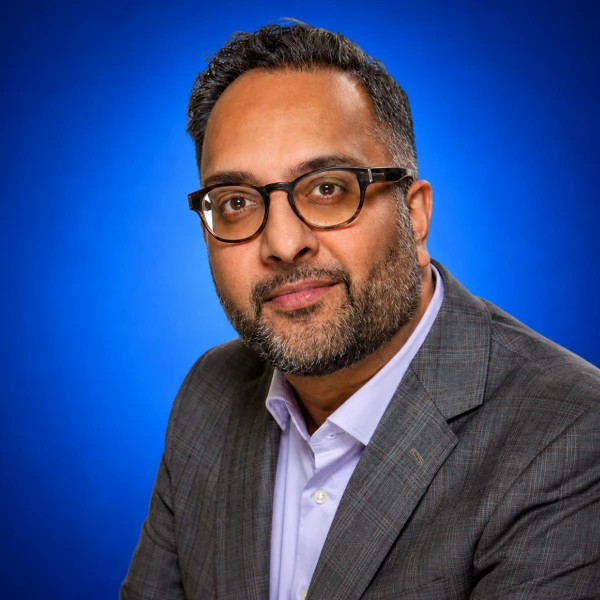 Professional headshot of a man with glasses in a gray suit and lavender shirt against a blue background.