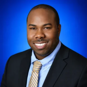 Smiling Black man in a navy suit, light blue shirt, and yellow striped tie, against a blue gradient backdrop (professional headshot).