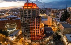Aerial view of a city center at dusk with illuminated streets and orange-lit circular rooftop on a tall glass tower in the foreground.