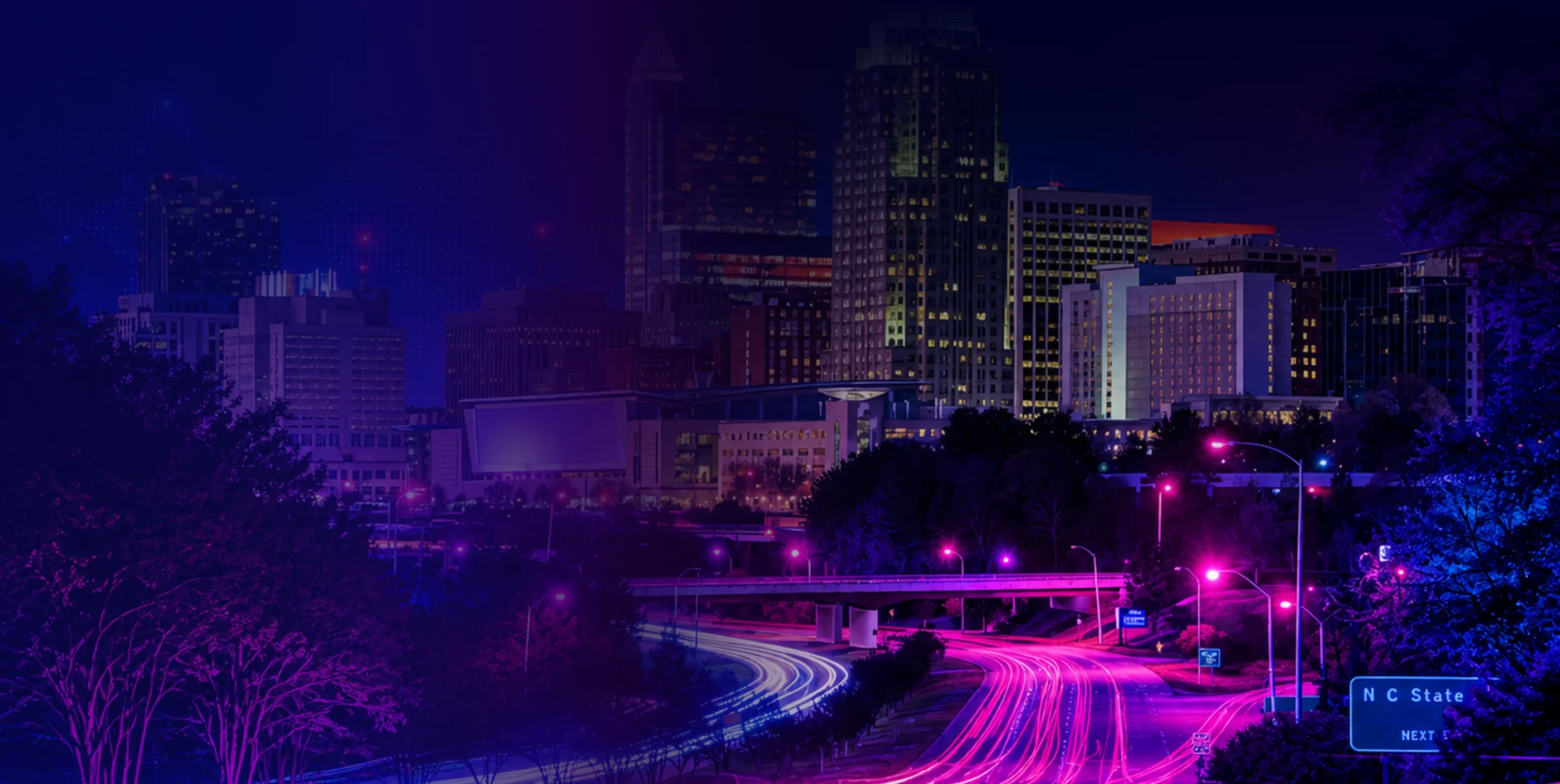 Nighttime city skyline bathed in blue-purple lights, with glowing highway light trails in the foreground and an NC State Next sign visible.