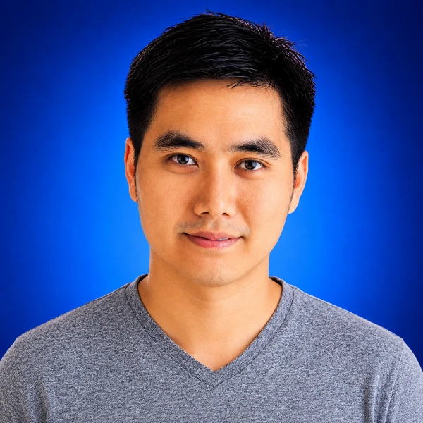 Portrait of a young man with short black hair wearing a gray V-neck shirt, against a blue studio backdrop.