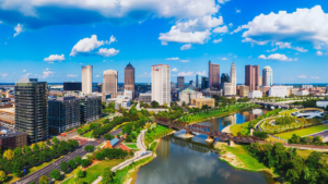 Aerial view of a modern city skyline by a winding river with several bridges and lush green parks under a bright blue sky.