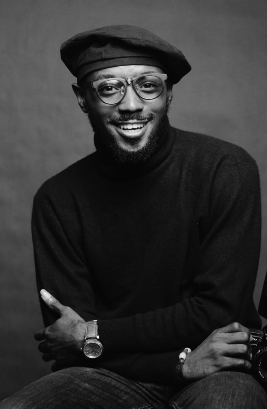 Black-and-white portrait of a smiling man wearing glasses and a hat, in a dark sweater, with a watch on his left wrist and a camera visible at the edge of the frame.