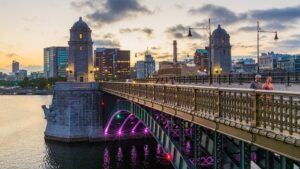 Evening view of a historic bridge with stone towers, ornate railing, and pedestrians, skyline in the background at sunset with purple-lit arches beneath the bridge.