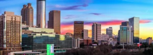 City skyline at sunset with tall glass buildings, a ferris wheel, and a colorful sky fading from blue to pink and orange.