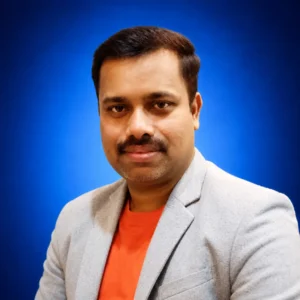 Anoop Karumambil Professional headshot of a man in a light gray blazer and orange shirt, smiling slightly against a blue gradient background.