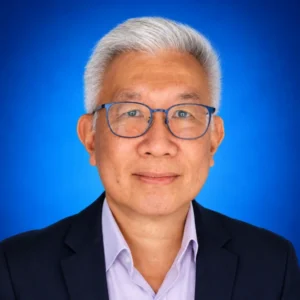 Portrait of a middle-aged Asian man with short gray hair and blue glasses, wearing a dark blazer and light purple shirt against a blue studio background, as a professional headshot.