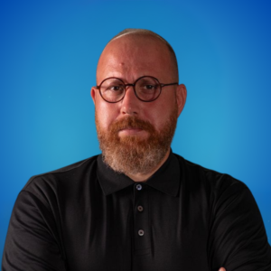 Portrait of a bald man with round glasses and a beard, wearing a black polo shirt, arms crossed against a blue gradient background.