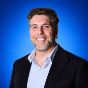 Todd Q. Chapman Professional headshot of a smiling man in a navy blazer and light blue shirt against a solid blue studio background.