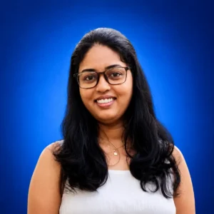 Divya-Marappan Smiling woman with dark hair and glasses against a blue studio background.