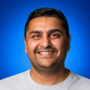 Namresh Saraiya Portrait of a smiling man with short dark hair and light stubble, wearing a gray shirt against a blue backdrop and studio lighting.