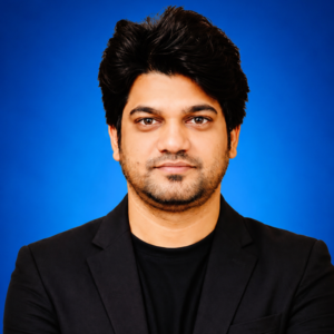 Professional head-and-shoulders portrait of a man with dark hair and stubble, wearing a black blazer against a blue background