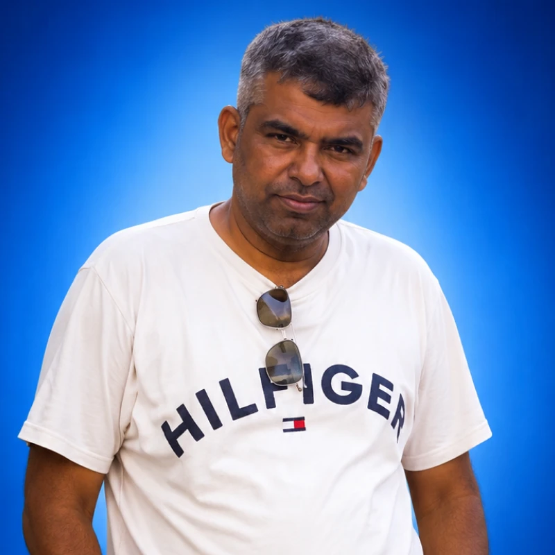 Man in a white Tommy Hilfiger T-shirt wearing sunglasses hanging from neckline, against a blue gradient backdrop.