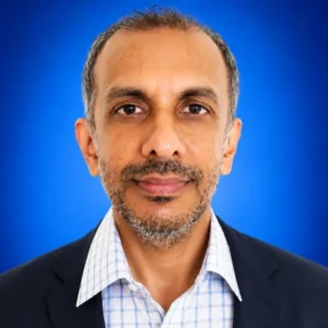 Professional headshot: middle-aged man in a dark blazer and light checkered shirt, facing the camera against a blue studio backdrop.