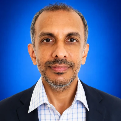 Professional headshot: middle-aged man in a dark blazer and light checkered shirt, facing the camera against a blue studio backdrop.