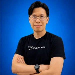 Portrait of a man with glasses wearing a black INVOLVE ASIA t-shirt, arms crossed against a blue background.