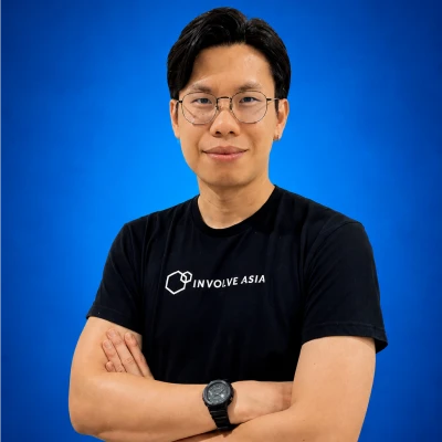 Portrait of a man with glasses wearing a black INVOLVE ASIA t-shirt, arms crossed against a blue background.