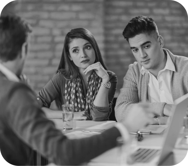 Three professionals sit at a table in a meeting, a woman gazing thoughtfully while a man to her right listens, a colleague facing away in the foreground.