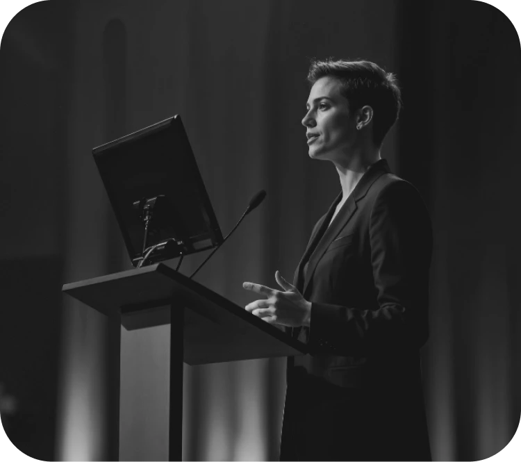 Woman in a blazer presenting at a lectern with a monitor and mic on stage in black and white; mid-speech gesture.