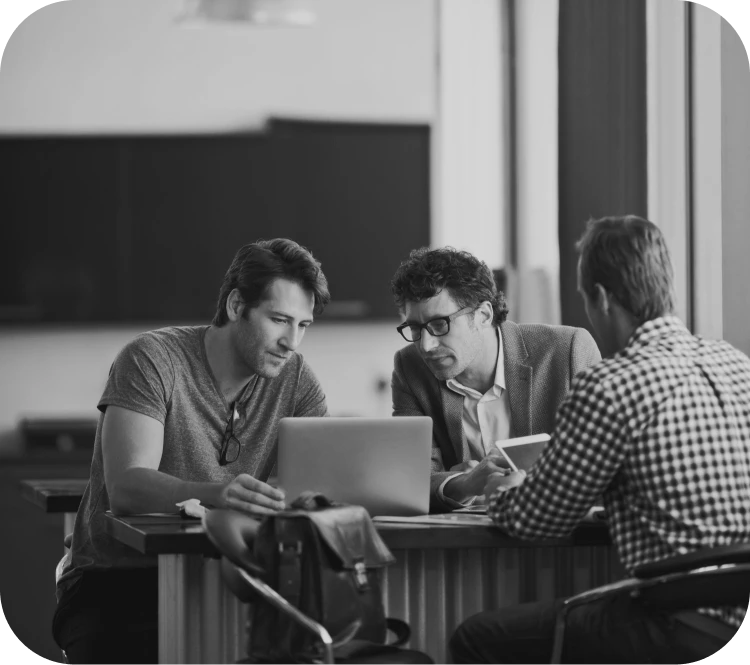 Three men collaborate at a counter with a laptop, discussing a project in a bright office.