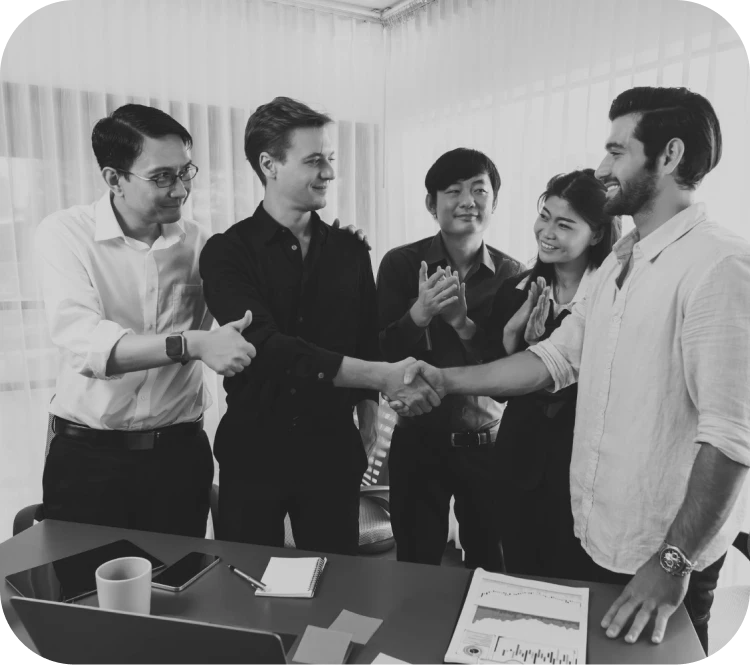 Five professionals in an office shake hands between two men while the others applaud, signaling a deal or agreement.