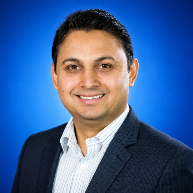 Professional headshot of a smiling man in a dark blazer and light striped shirt against a blue background, suitable for a corporate profile.