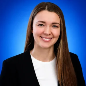 Professional headshot of a smiling woman with long brown hair, wearing a black blazer against a blue gradient background.
