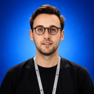 Andrew Knight Portrait of a man wearing round glasses and a black shirt against a blue gradient background, with a gray lanyard around his neck.