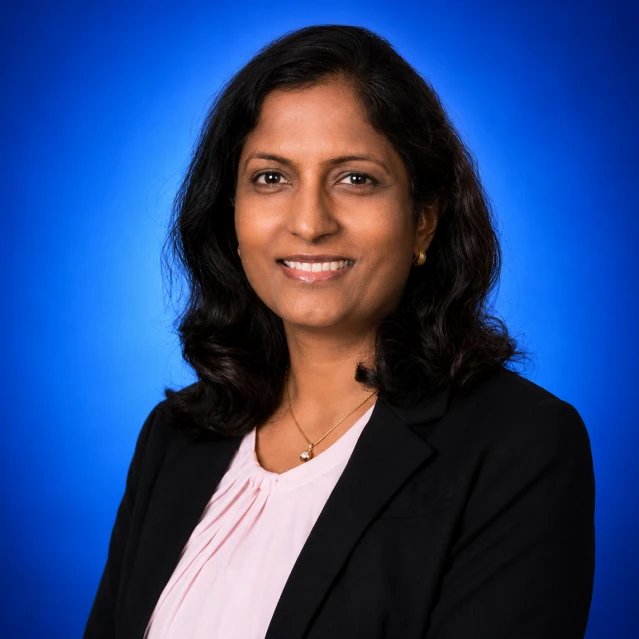 Professional headshot of a smiling woman with dark hair, wearing a black blazer and light pink blouse against a blue backdrop.