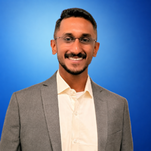 ChatGPT Image Jul 11, 2026, 01_33_16 PM Professional headshot of a smiling man in a gray suit and white shirt against a blue gradient backdrop.