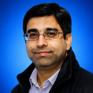 Head-and-shoulders portrait of a man wearing glasses and a dark jacket, posed against a blue background.