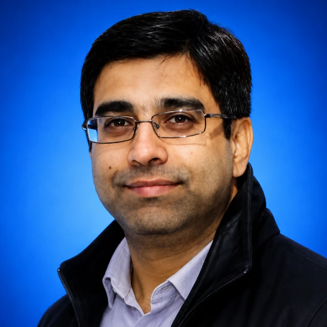 Head-and-shoulders portrait of a man wearing glasses and a dark jacket, posed against a blue background.