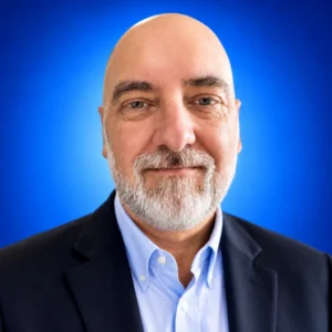 Smiling bald man with gray beard in a dark suit and light blue shirt against a blue gradient background.