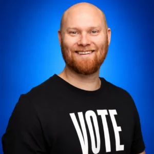 Smiling man with a red beard wearing a black T-shirt that says VOTE against a bright blue background.